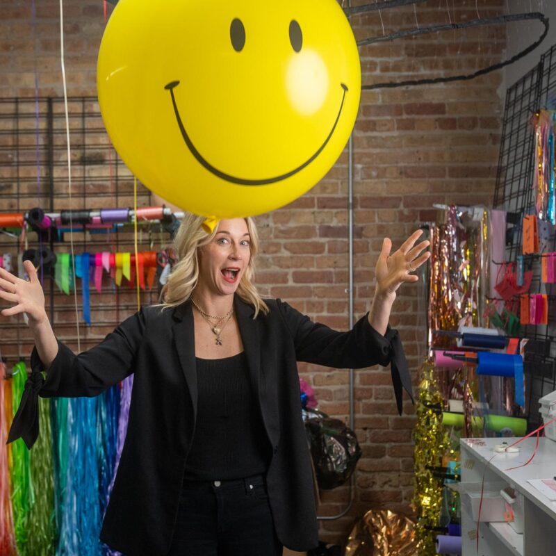 Elaine standing in her store, releasing a smiley face balloon.